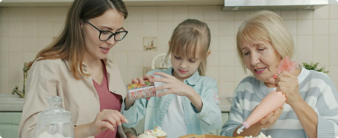 Drei Generationen von Frauen: eine Seniorin, eine Erwachsene und ein Mädchen backen gemeinsam Cup-Cakes in einer Küche. 