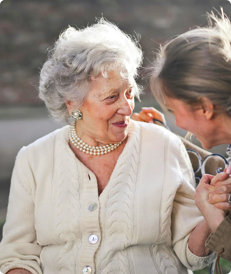 Eine Seniorin schaut glücklich eine ihr zugeneigte Frau an während sich beide an der Hand halten.