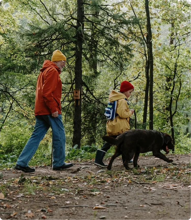 Ein Senioriger Mann mit Wanderstock und ein Junge spazieren gemeinsam mit einem Hund einen Waldweg entlang.