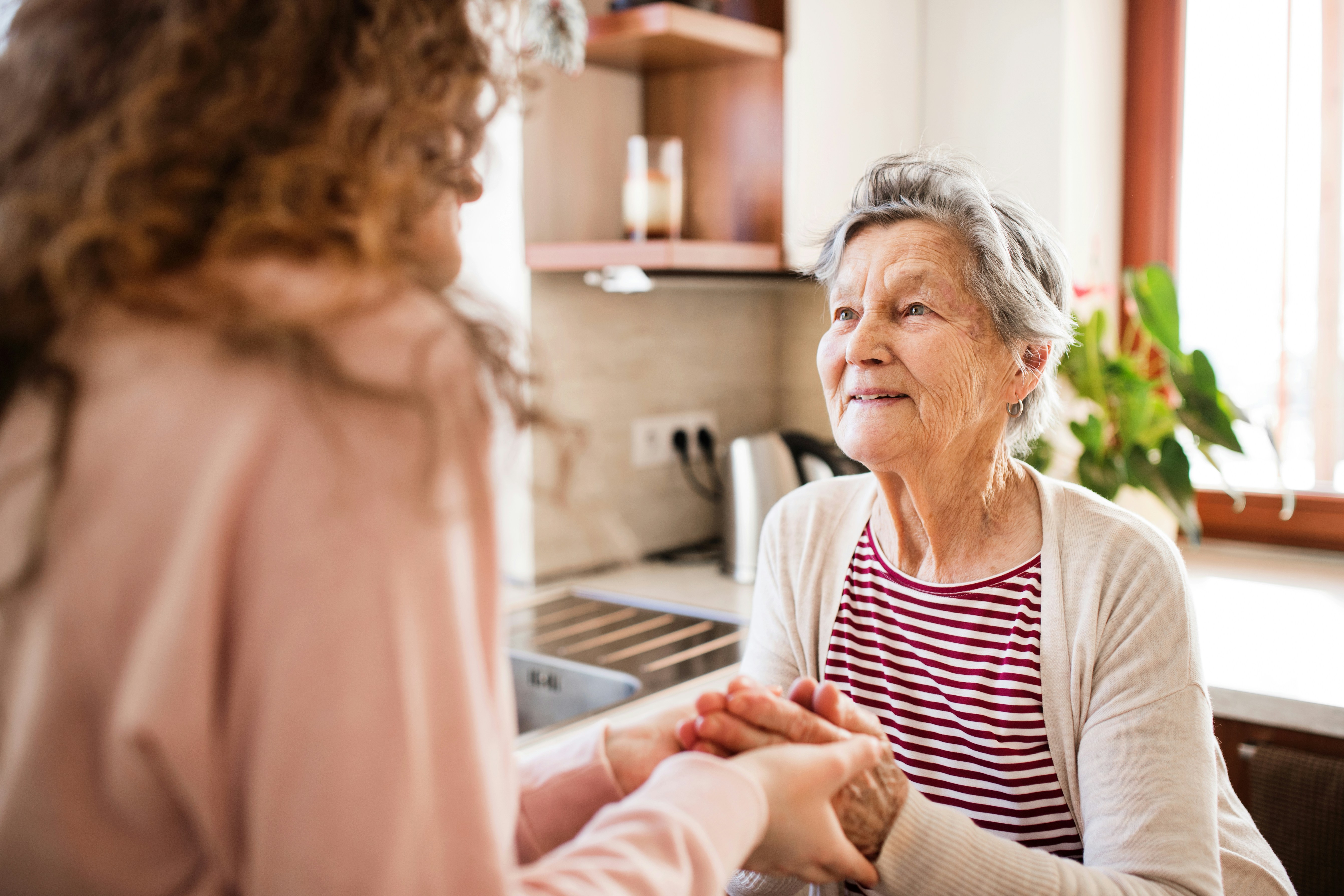 Eine junge Frau hält die gefalteten Hände einer sie anschauenden Seniorin in den eigenen Händen.